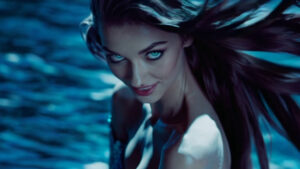 A mysterious woman with glowing aqua-blue eyes looks directly at the viewer, her dark hair flowing around her as she emerges from shimmering blue water under dramatic lighting.